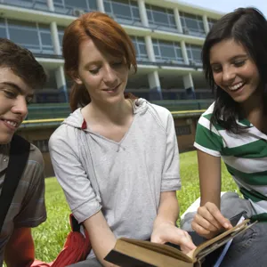 Tre ungdommer sitter på gressplenen utenfor en skole.