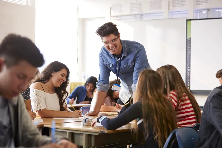 En ung, mannlig lærer smiler og lener seg på et bord over en gruppe elever ved pultene sine.